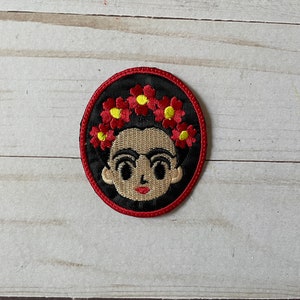 Puede incluir: Un parche redondo bordado con un retrato de dibujos animados de Frida Kahlo con una corona de flores rojas. El parche tiene un fondo negro y un borde rojo.