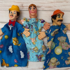 May include: Three hand puppets with different facial expressions and clothing. The puppet on the left is wearing a yellow hat and a blue and white patterned shirt. The puppet in the middle is wearing a blue and white patterned shirt with a star design. The puppet on the right is wearing an orange hat and a blue and white patterned shirt with a bear design.