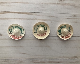 Può includere: Tre piccoli cappelli di paglia con le parole "Viva Mexico" e un design di cactus stampato su di essi.