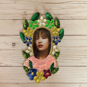 May include: A framed photo of a woman with dark hair and a pink shirt, surrounded by a floral design made of sequins and beads in shades of green, blue, yellow, white, and pink.