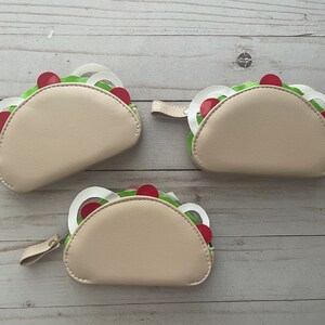 May include: Three taco-shaped zipper pouches made of beige faux leather. Each pouch features decorative cutouts representing taco fillings, including green lettuce, white onion rings, and red tomato circles, with a small beige pull tab.