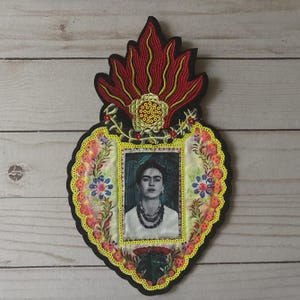 Puede incluir: Parche bordado con un retrato de Frida Kahlo dentro de un marco en forma de corazón. El marco está adornado con motivos florales y de llamas en rojo, amarillo y verde, sobre un fondo negro. El parche está diseñado para aplicarse en ropa o accesorios.