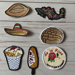 Pode incluir: Uma coleção de patches bordados com temas mexicanos. Inclui um chapéu mexicano, um mapa do México, uma concha, um copo de sorvete, uma espiga de milho no palito e um patch com o texto "de la Rosa".