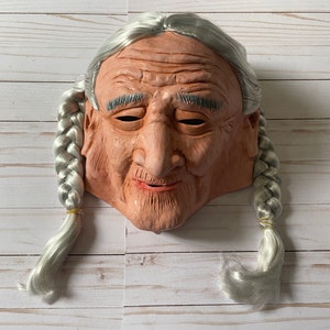 May include: A light brown latex mask of an elderly person with gray braided hair and a smiling expression.
