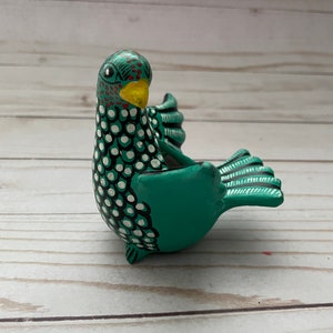 May include: A hand-painted ceramic bird figurine in teal, black, white, yellow, and red. The bird has a detailed pattern of white dots and black outlines on its body. The beak is yellow, and the head features red and black accents.