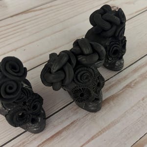 May include: Three black skull figurines with intricate details. Each skull has a unique design, including floral and braided elements. The skulls are arranged on a light-colored wooden surface, creating a striking contrast.