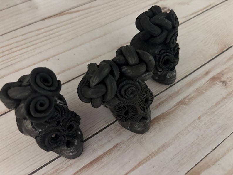 May include: Three black skull figurines with intricate details. Each skull has a unique design, including floral and braided elements. The skulls are arranged on a light-colored wooden surface, creating a striking contrast.