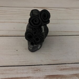 May include: A small, dark gray skull figurine adorned with black rose embellishments. The skull has a weathered appearance, with visible texture and subtle highlights. The roses are clustered on the forehead and around the eye sockets, creating a gothic or Day of the Dead theme.