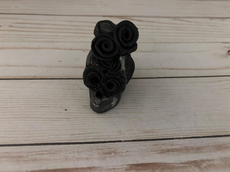 May include: A small, dark gray skull figurine adorned with black rose embellishments. The skull has a weathered appearance, with visible texture and subtle highlights. The roses are clustered on the forehead and around the eye sockets, creating a gothic or Day of the Dead theme.