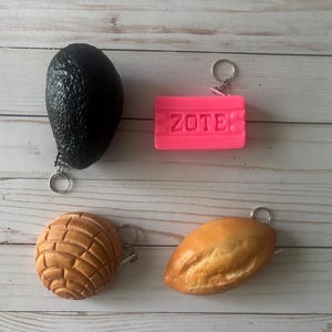 Peut inclure: Quatre porte-clés sont présentés : un avocat, un pain de savon rose avec le texte "ZOTE", une pâtisserie concha et un petit pain. Chaque porte-clés a un anneau argenté. L'avocat est noir, le savon est rose vif, la concha est beige et le petit pain est brun doré.