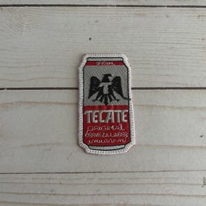 May include: Embroidered patch featuring a stylized design of a beer can. The patch has a red border and a gray background with the word "TECATE" in red. A black eagle emblem is centered above the text.