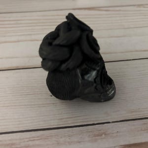May include: A black, sculpted figurine with a textured surface. The figurine has a rounded base and a top section with a series of curved, layered elements. The object is set against a light wood-grain background.