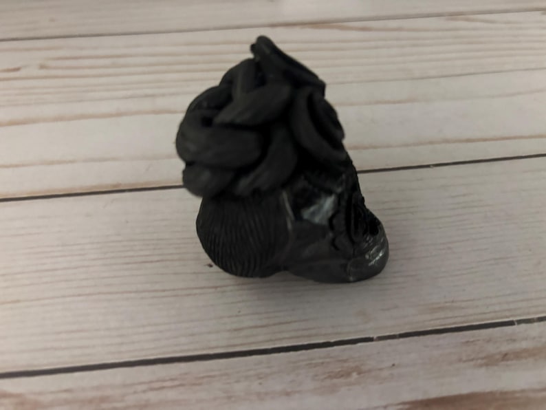 May include: A black, sculpted figurine with a textured surface. The figurine has a rounded base and a top section with a series of curved, layered elements. The object is set against a light wood-grain background.