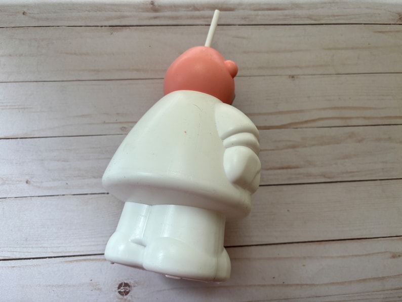 Peut inclure: Une figurine en plastique blanche repr&eacute;sentant une personne avec une t&ecirc;te rose et une paille qui sort du haut. La figurine porte un manteau blanc et a un bras blanc avec un gant blanc.