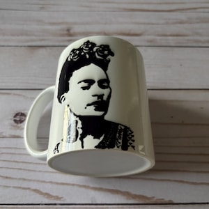 May include: Cream ceramic mug with a black and white portrait of a woman wearing a floral headpiece. The portrait is a stylized illustration. The mug has a white handle and a wide base.