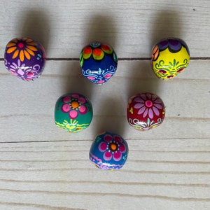 May include: Six hand-painted, oval beads with floral designs. The beads are painted in various colours, including purple, blue, yellow, green, and red. Each bead features a different floral pattern, with pink, orange, and white accents. The beads are arranged on a light wood surface.