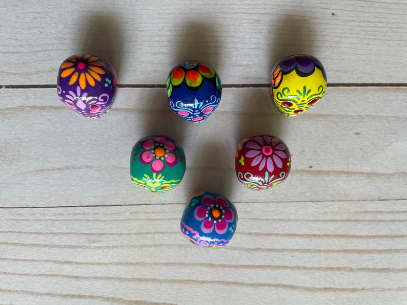 May include: Six hand-painted, oval beads with floral designs. The beads are painted in various colours, including purple, blue, yellow, green, and red. Each bead features a different floral pattern, with pink, orange, and white accents. The beads are arranged on a light wood surface.
