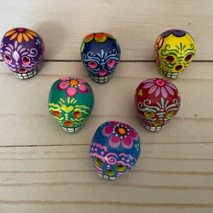May include: Six hand-painted Day of the Dead sugar skull figurines in various colours, including purple, blue, yellow, green, and red. Each skull features intricate floral and decorative designs, with white teeth and eye sockets.