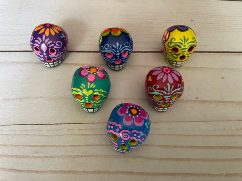 May include: Six hand-painted Day of the Dead sugar skull figurines in various colours, including purple, blue, yellow, green, and red. Each skull features intricate floral and decorative designs, with white teeth and eye sockets.