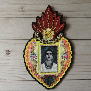 Może przedstawiać: Naszywkę w kształcie serca, haftowana, z obrazkiem Fridy Kahlo w środku. Serce otoczone jest kolorowymi kwiatami i wzorem czerwonego płomienia.