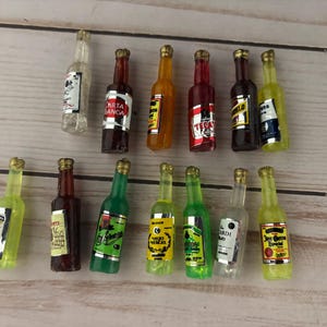 Pode incluir: Miniaturas de garrafas de licor em várias cores, incluindo transparente, vermelho, amarelo e verde. Algumas garrafas têm tampas douradas e rótulos com texto. Dispostas sobre uma superfície de madeira clara.
