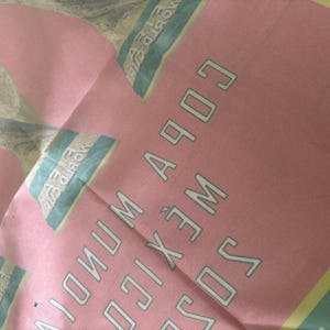 May include: Pink fabric with text that reads "COPA MUNDIAL MEXICO 1970". The text is in a white, outlined font. The fabric also has a light blue and yellow design.