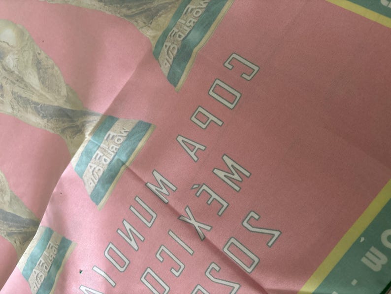 May include: Pink fabric with text that reads "COPA MUNDIAL MEXICO 1970". The text is in a white, outlined font. The fabric also has a light blue and yellow design.
