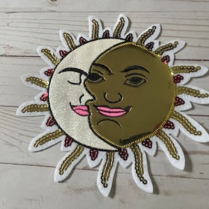 May include: A white and gold sun and moon patch with pink lips and eyes. The sun and moon are kissing. The patch is made of felt and sequins.