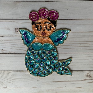 May include: A blue and purple sequined mermaid with a pink flower crown and black hair. The mermaid has a gold outline and is wearing glasses.