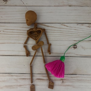 May include: A brown articulated skeleton doll with a pink tassel broom. The doll is made of a light brown material and has movable joints. The broom is made of pink paper and has a green handle.