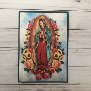 Peut inclure: Une image colorée de la Vierge Marie, représentée dans une robe bleue et dorée, avec une couronne d'étoiles. Elle est entourée de tournesols, de roses et d'autres fleurs. L'image est sur un fond blanc.