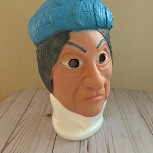 Puede incluir: Una máscara de plástico azul y gris con una expresión severa. La máscara tiene un sombrero azul y está sobre un soporte blanco.