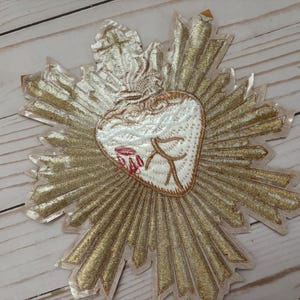 Puede incluir: Un parche bordado en dorado con un coraz&oacute;n blanco en el centro. El coraz&oacute;n tiene un dise&ntilde;o rojo y blanco y est&aacute; rodeado por un halo dorado.