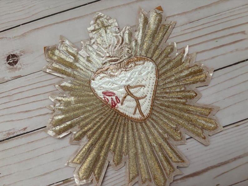 Puede incluir: Un parche bordado en dorado con un coraz&oacute;n blanco en el centro. El coraz&oacute;n tiene un dise&ntilde;o rojo y blanco y est&aacute; rodeado por un halo dorado.