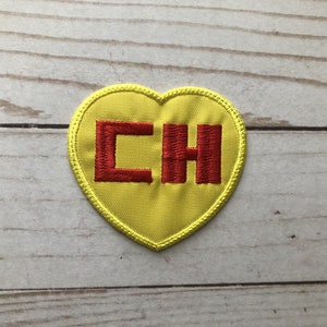 Pode incluir: Patch bordado amarelo em forma de coração com as letras vermelhas "CH". O patch tem uma borda amarela e está sobre um fundo claro de grão de madeira. O patch é provavelmente para roupas ou acessórios.