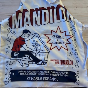 Può includere: Grembiule bianco con la parola "MANDILON" in grandi lettere color marrone. Un uomo dei cartoni animati con una camicia rossa è raffigurato al lavoro. Il grembiule ha una tasca con testo in spagnolo, tra cui "OBEDIENTE Y CONFIABLE".