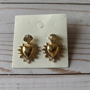 May include: Gold-colored heart-shaped earrings with a sunburst design. The earrings have a shell-like detail at the top and are displayed on a white card. The earrings are a jewelry item.