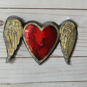 May include: A sequined patch featuring a red heart with gold wings. The heart is made of red sequins and outlined in gold. The wings are gold sequins on a gray fabric backing. This patch is suitable for clothing or accessories.