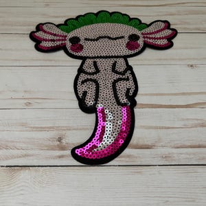 Puede incluir: Un ajolote rosa y verde con un diseño de lentejuelas brillantes. El ajolote es un tipo de salamandra nativa de México.