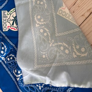 May include: A blue and light blue bandana with a paisley design and the word "MEXICO" printed on it. The bandana also features the Mexican national emblem. The fabric is folded, revealing both sides of the bandana.