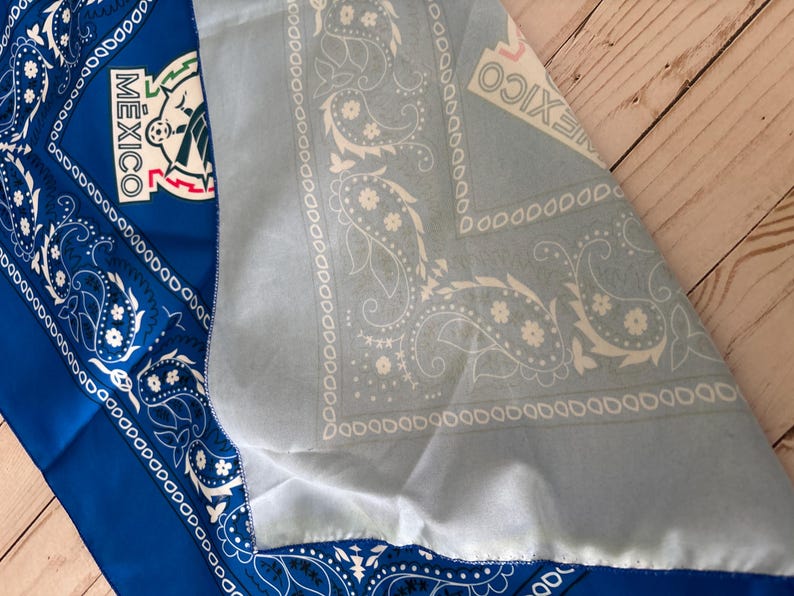 May include: A blue and light blue bandana with a paisley design and the word "MEXICO" printed on it. The bandana also features the Mexican national emblem. The fabric is folded, revealing both sides of the bandana.