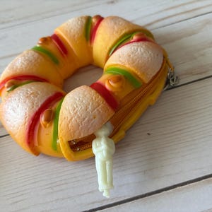 Puede incluir: Un monedero novedoso con forma de roscón de Reyes, con detalles de glaseado amarillo, rojo y verde. La cremallera está abierta, revelando una pequeña figura de plástico. El monedero está sobre una superficie de madera clara.