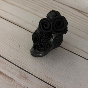 May include: A black skull-shaped decorative object with two rose-shaped embellishments on top. The skull has intricate floral patterns and is set against a light wood grain background.