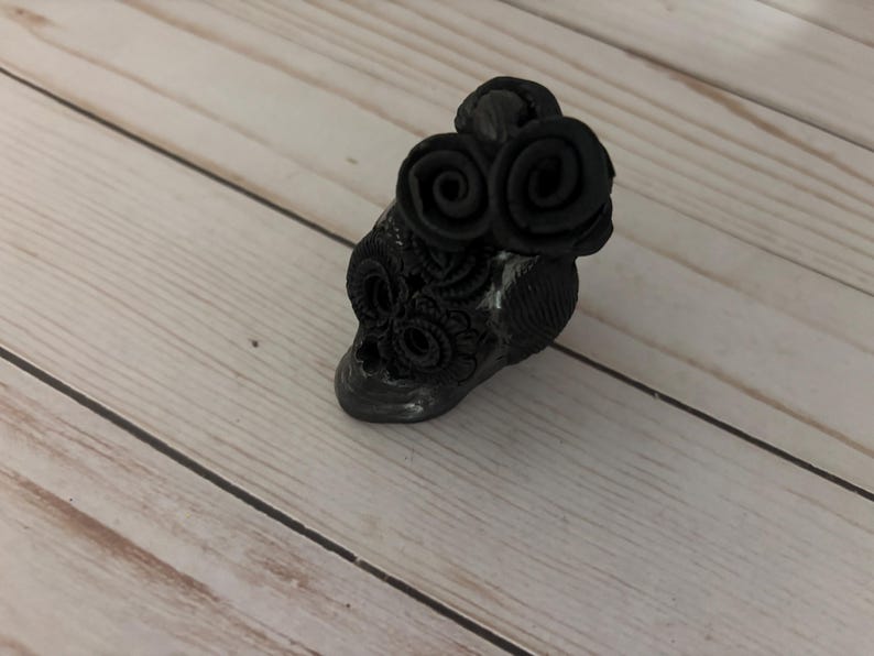 May include: A black skull-shaped decorative object with two rose-shaped embellishments on top. The skull has intricate floral patterns and is set against a light wood grain background.