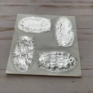 May include: Four silver foil stickers featuring an image of the Virgin Mary. The stickers are on a white sheet of paper.