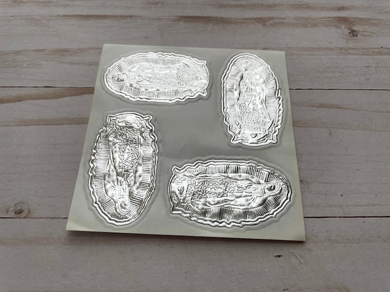 May include: Four silver foil stickers featuring an image of the Virgin Mary. The stickers are on a white sheet of paper.