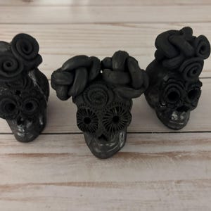 May include: Three black skull figurines with intricate floral and braided hair details. The skulls have detailed eye sockets and are arranged on a light wood surface. The figurines are a dark gray color.