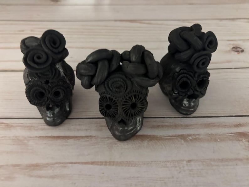 May include: Three black skull figurines with intricate floral and braided hair details. The skulls have detailed eye sockets and are arranged on a light wood surface. The figurines are a dark gray color.