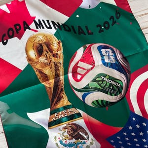 May include: A decorative scarf featuring the FIFA World Cup trophy and a soccer ball. The scarf has the text "COPA MUNDIAL 2026" and includes flags of the United States, Mexico, and Canada. The soccer ball has the word "TRIONDA" on it.