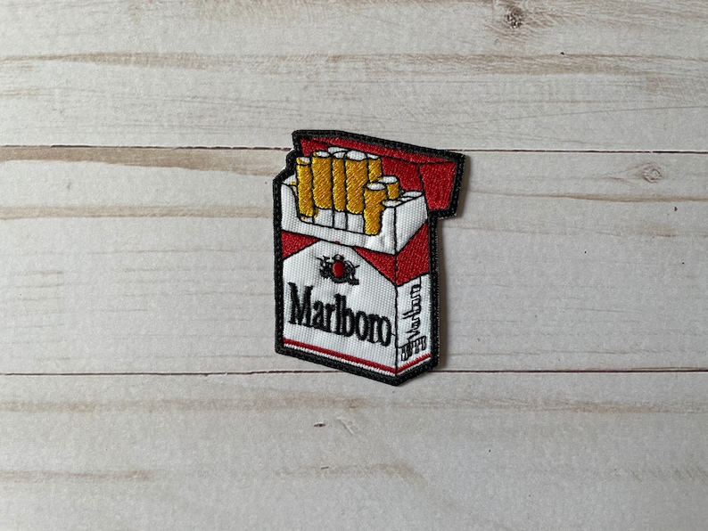 May include: Embroidered patch featuring a pack of Marlboro cigarettes. The patch has a white background with red accents and the Marlboro logo in black. The cigarettes are yellow and white.